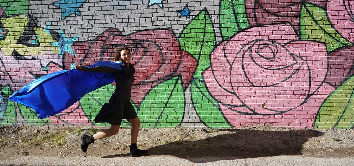 Woman running with blue cape flying in front of graffiti wall